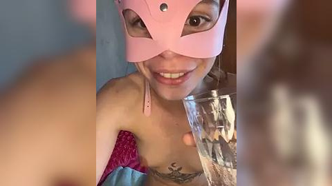 Media: Video of a smiling woman with fair skin, wearing a pink mask, a pink top, and blue fabric, with a tattoo on her chest.