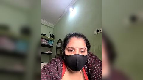 Media: Video of a woman with medium skin tone, wearing a black face mask, black blouse, and a dark patterned jacket, seated in a green-painted room with a white ceiling and shelves filled with items.