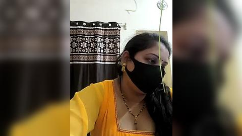 Media: Video of an Indian woman with medium skin tone, wearing a yellow saree, black mask, and gold earrings, indoors with a patterned curtain and a mirror.