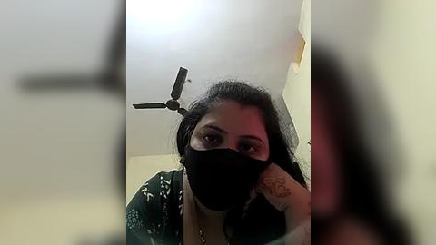 Media: Video of a woman with dark hair and medium skin tone wearing a black face mask, black top, and white floral patterned cardigan, standing in a room with a ceiling fan and a yellow wall.