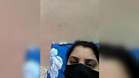 Media: Video of a woman lying on a blue pillow with white flower pattern, wearing a black mask, with visible nose and mouth. Background shows a beige wall with minor scratches.