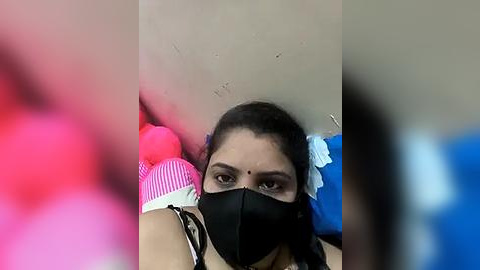 Media: A video of a woman with brown skin, wearing a black mask, black top, and pink striped sleeve, holding a stuffed pink toy. The background is blurred, with a beige wall.