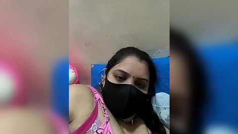 Media: Video of a South Asian woman with medium brown skin, wearing a black face mask and a pink top, lying on a blue bed, with a beige wall background.