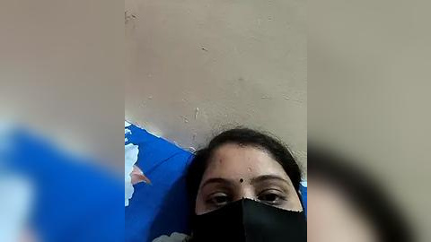 Media: Video of a woman with dark hair and a black mask, lying on a blue tarp against a beige, peeling wall, looking directly at the camera.