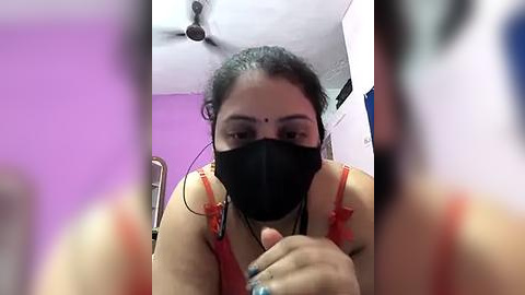 Media: Video of a South Asian woman with dark hair and medium skin tone, wearing a black face mask and a red bra, indoors with purple and white walls, ceiling fan, and blurred background.