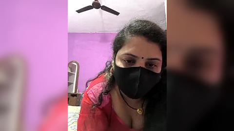 Media: Video of a woman with dark skin and curly hair, wearing a red blouse and black mask, sitting on a bed in a purple room with a ceiling fan.