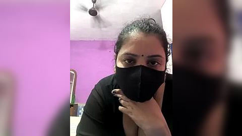 Media: A video shows a South Asian woman with medium skin tone, dark hair tied back, wearing a black face mask, black top, and sitting in a room with purple walls and a ceiling fan.