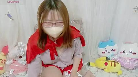 Media: Video of a young Asian woman in a red cape, wearing a face mask, red shorts, and a gray top. She's seated on a white bed with Pikachu and Hello Kitty plushies in the background.