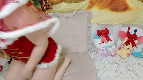 Media: A video shows a young girl in a red Santa dress, with a blurred face, standing near a bed with white sheets and pillows. A teddy bear and plush toys are beside the bed.