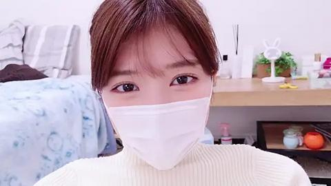 Media: Video of a young Asian woman with light skin and short brown hair, wearing a white face mask, a cream-colored ribbed sweater, and sitting in a modern bedroom with a bed, dresser, and various household items.