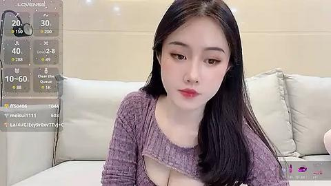 Media: A video of an East Asian woman with long black hair, fair skin, and red lipstick, wearing a purple knit top with a cut-out, sitting on a white couch in a modern living room.