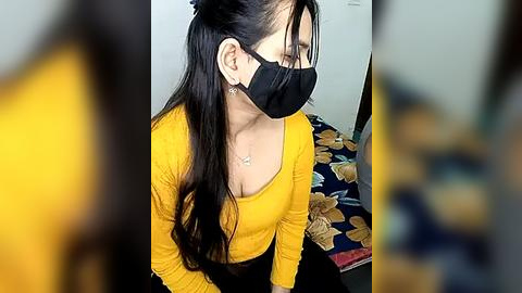 Media: Video of a woman with long black hair, wearing a yellow long-sleeved top, black face mask, and earrings, sitting on a bed with floral sheets.