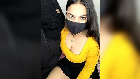 Media: A video of a young woman with long black hair and light skin, wearing a yellow top and black face mask, sitting on a black couch.