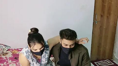 Media: Video of an Indian woman with dark hair in a bun, wearing a floral blouse and face mask, sitting next to a masked man with short dark hair, in a simple bedroom with a wooden door.