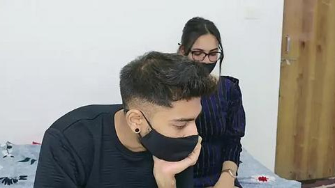 Media: Video of a man with short, dark hair, wearing a black face mask and black long-sleeve shirt, sitting on a bed with floral-patterned sheets. A woman with long dark hair and glasses stands behind him, wearing a dark patterned blouse.