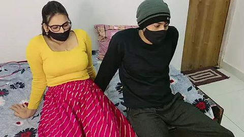 Media: Video of two people, a woman in yellow top and red skirt, and a man in black shirt, seated on a bed with floral sheets in a simple room.