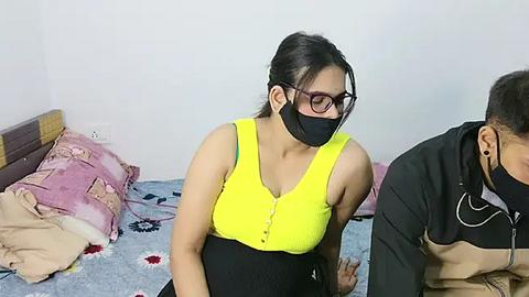 Media: Video of a young woman with a medium complexion, wearing a yellow top, black mask, and glasses, seated on a bed with a pink and blue blanket, surrounded by a man in a black jacket and another person partially visible.