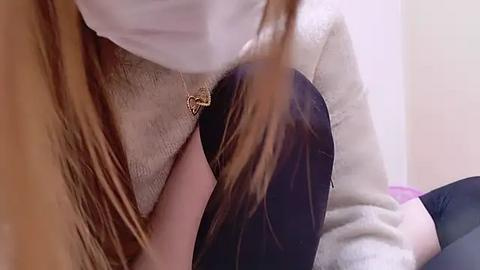 Media: Video of a person with long, light brown hair, wearing a white face mask, beige sweater, and black leggings, seated on a beige couch in a softly lit room.