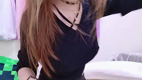 Media: Video of a woman with long, straight, light brown hair, wearing a black, ribbed knit top and a choker necklace, standing in a brightly lit room with a bed and green and white bedding in the background.