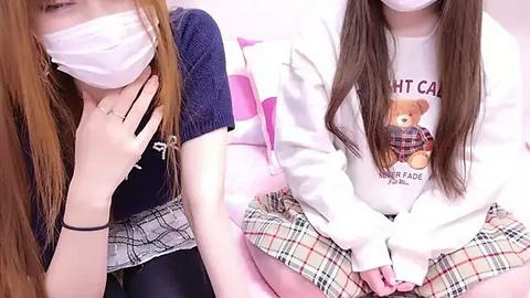 Media: Video of two Asian women wearing face masks, one in a dark sweater and plaid skirt, the other in a white hoodie with a teddy bear graphic and plaid skirt, seated on pink bedding.