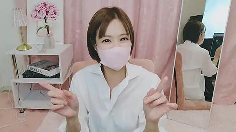 Media: A video of an East Asian woman with shoulder-length brown hair, wearing a white blouse and a pink surgical mask, sitting in a pink chair, gesturing with both hands, in a pink room with a white shelf and flowers.