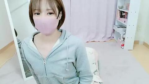 Media: A young woman with short, light brown hair, wearing a light blue hoodie, a pink face mask, and standing in front of a mirror in a softly lit room with white walls and a pastel pink curtain.