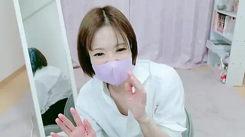 Media: Video of a young Asian woman with short brown hair, wearing a white lab coat, and a light purple face mask, sitting at a desk in a clinical room with light pink curtains and white cabinets.