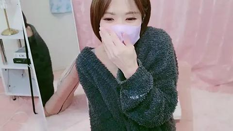 Media: Video of an Asian woman with short brown hair, wearing a dark gray fuzzy sweater and a purple mask, kneeling on pink carpet in a room with light pink curtains and a white bookshelf.