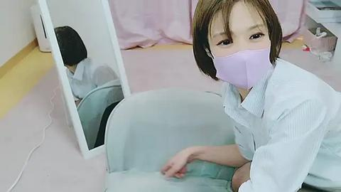 Media: Video of an Asian woman with short brown hair, wearing a light blue striped shirt and purple face mask, kneeling in front of a mirror, reflecting her image.