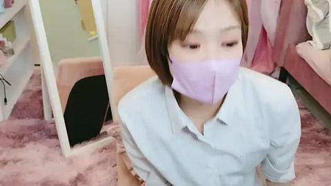 Media: A video of an East Asian woman with light skin, short brown hair, and a lavender face mask, sitting on a pink shag rug in a cluttered, pastel-colored room.