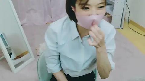 Media: A video of an Asian woman in a light blue shirt, wearing a pink face mask, covering her mouth, standing in a pink-tiled room with a mirror and a white shelf.