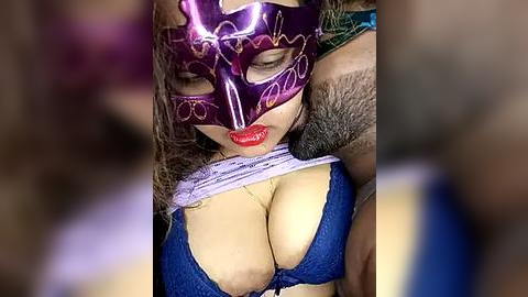 Media: Video of a woman with a purple masquerade mask covering her eyes, red lips, large breasts in a blue lace bra, and a furry arm behind her.