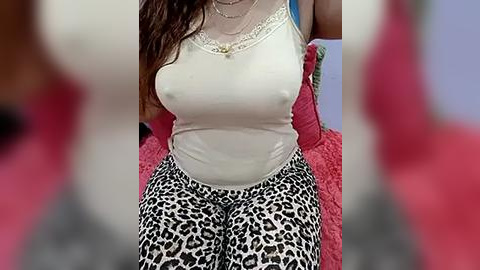 Media: Video of a woman with medium brown skin, wearing a white tank top and leopard-print pants, sitting on a red couch. She has long, wavy brown hair and is wearing multiple necklaces.