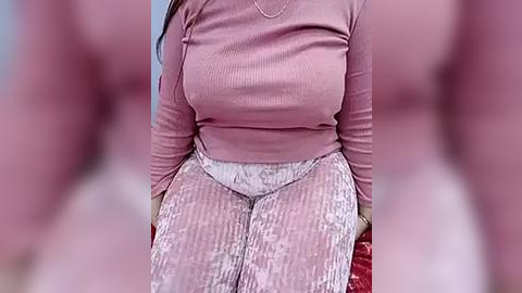 Media: Video of a woman sitting, wearing a pink ribbed sweater and white patterned leggings, with a blurred background.