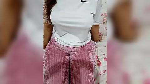 Media: A video of a woman with medium skin tone wearing a white T-shirt and pink, striped, high-waisted pants. The background features a white wall with pink floral patterns and blurred people.