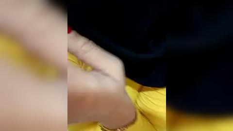 Media: A close-up video of a person's hand and forearm, partially obscured by a black sleeve and a yellow shirt, with a blurred background.