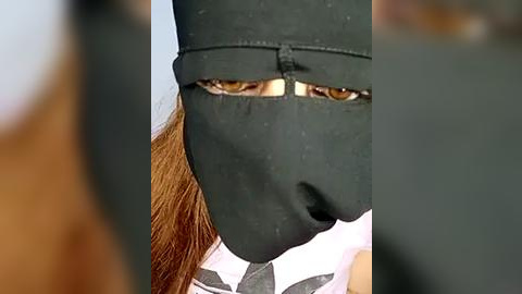 Media: Video of a person wearing a black balaclava with slit eyeholes, partially obscuring their face. Their long, reddish-brown hair peeks out from under the mask. The background is blurred, with hints of neutral colors.