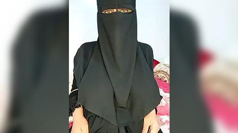 Media: Video of a woman in a black niqab, with a white headscarf partially visible, standing in a room with pink and white bedding, blurred background.