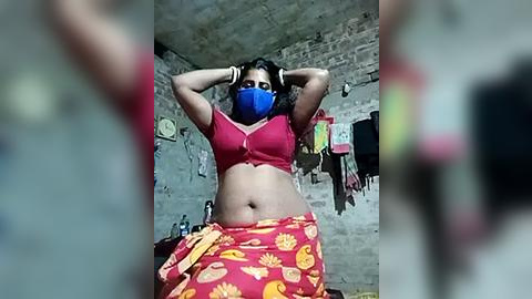 Media: A video of a South Asian woman with medium skin tone, wearing a red bra, blue mask, and a red saree with yellow sunflower pattern, standing in a dimly lit, rustic room with a brick wall.