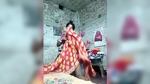 Media: Video of a woman in a red sari with yellow polka dots, standing in a simple, dimly lit room with white walls and a bed covered with colorful blankets.