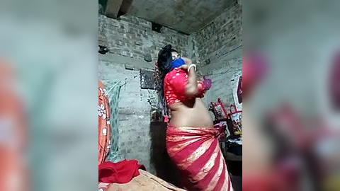 Media: Video of a woman with dark skin, wearing a red and gold sari, gagged with a blue cloth, tied to a brick wall in a dimly lit, rustic room.