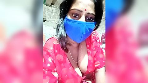 Media: Video of a South Asian woman with medium skin tone and medium-sized breasts wearing a bright pink sari with a deep neckline, a blue mask covering her nose and mouth, and a red bindi on her forehead. The background is blurred, suggesting an outdoor setting.