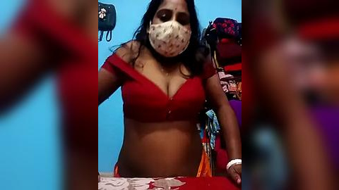 Media: A video of a South Asian woman with dark skin, wearing a red off-shoulder top and a face mask, standing in a cluttered room with blue walls and a red desk.