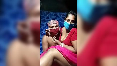 Media: Video of two women in masks and colorful sarees, one topless, in a blue patterned room; blurry blue background.