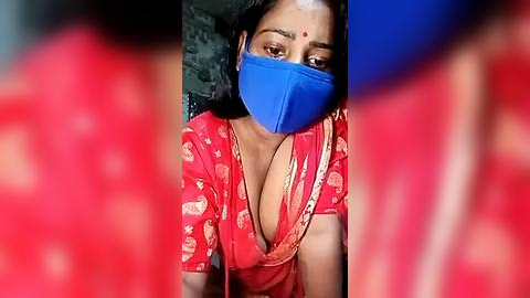 Media: A video shows a woman with medium brown skin, wearing a blue mask and a red saree with a deep neckline, revealing cleavage, against a blurred background.