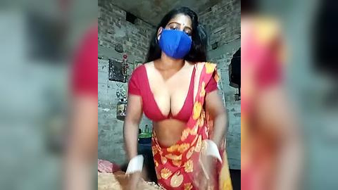 Media: Video of a South Asian woman with medium skin tone, wearing a red blouse, blue mask, and yellow sari, standing in a dimly lit, brick-walled room with posters on the wall.