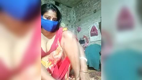 Media: Video of a woman in a red sari with a blue face mask, hands bound, in a dimly lit, cluttered room with posters and a bed.