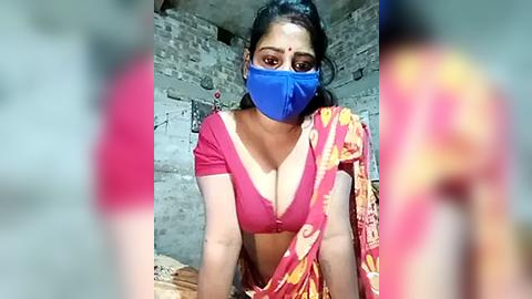 Media: Video of an Indian woman in a red blouse, blue face mask, and colorful sari, kneeling in a rustic, brick-walled room, her face partially blurred.