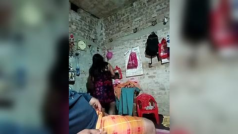 Media: A video of a woman in a purple floral dress hanging clothes in a rustic, brick-walled room with a blue plastic bag, red chair, and various personal items.