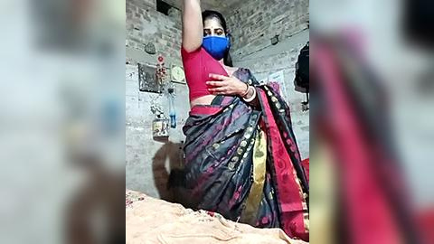 Media: Video of a South Asian woman in a red top and blue mask, with a multicolored sari, raising her arms, in a dimly lit, rustic room with exposed brick walls and personal items.